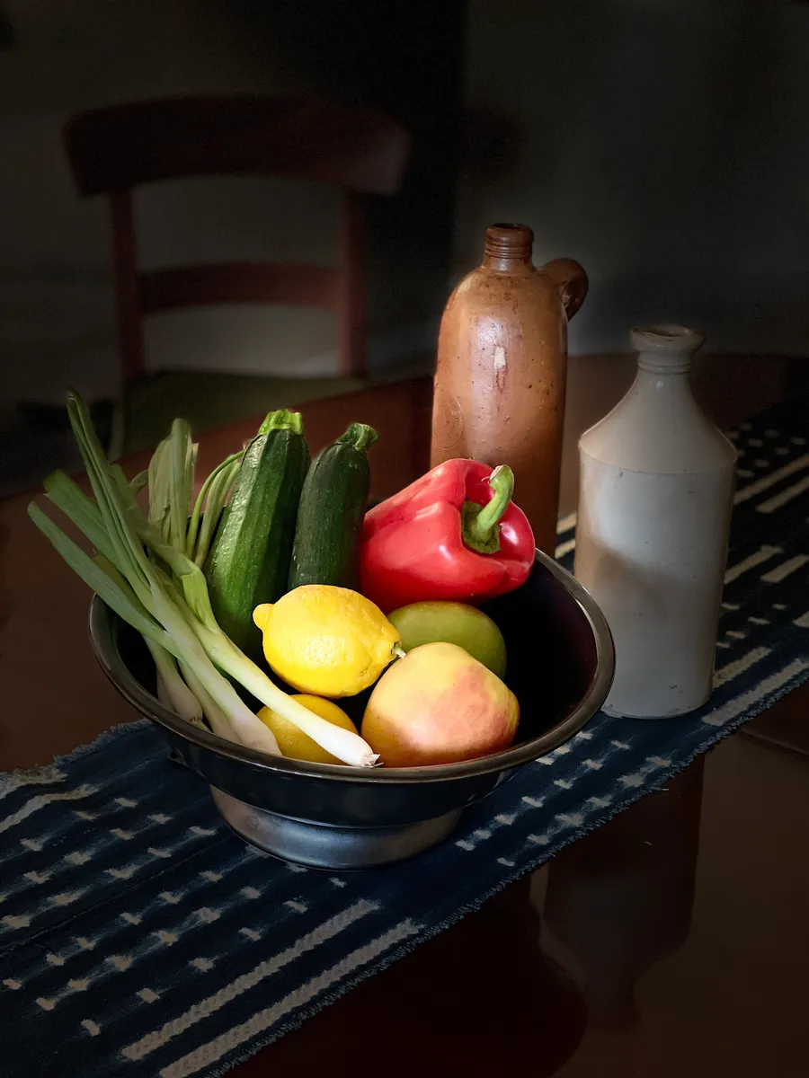 A dark bowl of lemons, a pear, a red pepper, zucchini and spring onions beside stoneware bottles, photograph by Esther Piaskowski