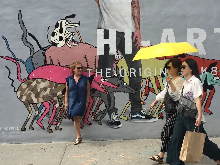 A woman in a blue dress stands against a street mural of cartoon animals while two women pass under a yellow umbrella, photograph by Esther Piaskowski